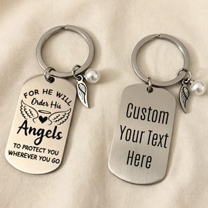 Op de afbeelding: Twee zilverkleurige metalen sleutelhangers in de vorm van dogtags met ringen. Eén hanger heeft gegraveerde engelenvleugels, een hartje en de tekst "FOR HE WILL ORDER HIS ANGELS TO PROTECT YOU WHEREVER YOU GO". De andere toont "CUSTOM YOUR TEXT HERE".