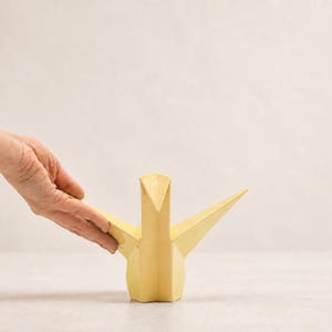 Op de afbeelding: Een lichtgeel keramieken sculptuur in de vorm van een origami kraanvogel. Het vogelbeeldje heeft scherpe, hoekige vouwen en geometrische vlakken die het uiterlijk van gevouwen papier nabootsen, met een spitse snavel, uitgestrekte vleugels en een stabiele voet.