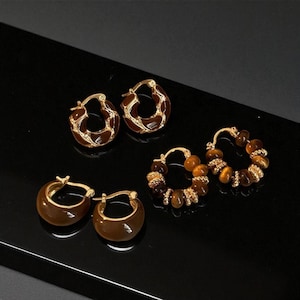 Peut inclure: Quatre paires de boucles d'oreilles créoles dorées. Deux paires présentent un motif en émail marron, et les deux autres paires sont ornées de perles marron. Les boucles d'oreilles sont présentées sur une surface noire.