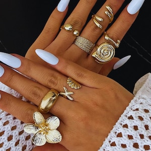 Può includere: Una collezione di anelli color oro in vari design, tra cui un fiore, una conchiglia, una stella marina e una spirale. Gli anelli sono mostrati su dita con unghie lunghe e azzurre. I gioielli sono su sfondo scuro.