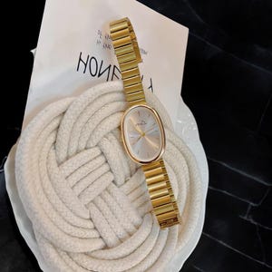 May include: Gold-coloured wristwatch with an oval face and a matching gold band. The watch has a light-coloured face with a small logo. The watch is displayed on a white surface with a rope design and a piece of paper with the word "HOME" printed on it.