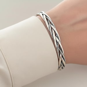 Op de afbeelding: Een zilveren armband met een ontwerp van meerdere strengen, bestaande uit een combinatie van gladde gepolijste metalen draden en gedraaide touwachtige strengen die in elkaar verweven zijn voor een complexe, gelaagde uitstraling.