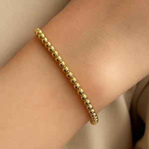 Könnte beinhalten: Ein goldfarbenes Perlenarmband bestehend aus einer einzelnen Reihe kleiner, gleichmäßiger, polierter kugelförmiger Perlen. Das Armband weist ein durchgehendes Design aus metallisch goldenen Kugeln auf, die zu einem einfachen, kreisförmigen Armschmuck verbunden sind.