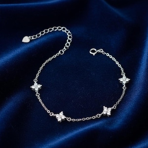 Peut inclure: Un bracelet en argent délicat avec quatre breloques en forme de fleur, chacune ornée de petites pierres scintillantes. Le bracelet est doté d'une chaîne réglable et d'un détail en forme de cœur à l'extrémité. Le bijou est présenté sur une surface en satin bleu foncé.