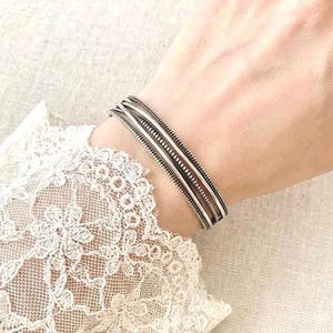 Könnte beinhalten: Ein silberner Armreif bestehend aus mehreren parallelen Bändern mit einer texturierten, geriffelten Oberfläche. Das Armband wird am Handgelenk getragen, das teilweise von einem weißen Spitzenärmel mit Blumenmuster bedeckt ist, was den Glanz betont.