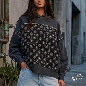 May include: A dark grey hoodie with a patterned front panel. The panel features a repeating floral design in shades of pink and white on a black background. The hoodie has a drawstring hood and long sleeves.