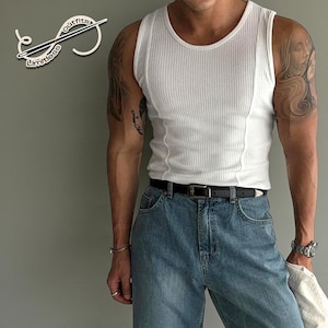 Puede incluir: Camiseta sin mangas blanca acanalada con cuello redondo y cinturón negro, combinada con vaqueros azules. La camiseta sin mangas tiene una textura acanalada vertical. Los vaqueros son de mezclilla de lavado claro. El logo "Limitless Outfits" está arriba a la izquierda.