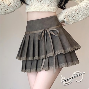 May include: A two-tiered, olive-green mini skirt with a pleated design. The skirt features a ribbon tie at the waist and delicate lace trim along the hemline. The skirt is part of the "Limitless outfits" collection.