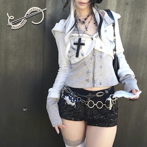 May include: A light gray and white jacket with a black cross pendant, paired with black sequin shorts. The outfit includes a silver chain belt and a gray top. The image has the text "Limitless Outfits".