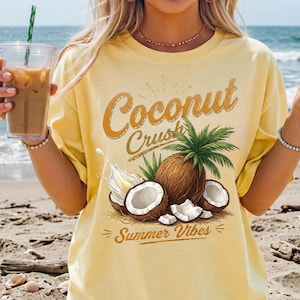 May include: A pale yellow t-shirt featuring a graphic print of coconuts, palm leaves, and a splash of liquid. The text reads Coconut Crush and Summer Vibes in a stylized brown font. The design is centered on the front of the short-sleeved garment.