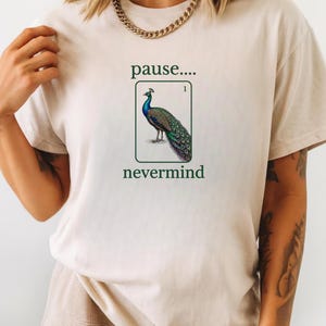 May include: A beige short-sleeve t-shirt featuring a graphic of a peacock inside a rectangular frame. Above the frame is the text "pause...." and below is the text "nevermind". The peacock is depicted in shades of blue, green, and brown.