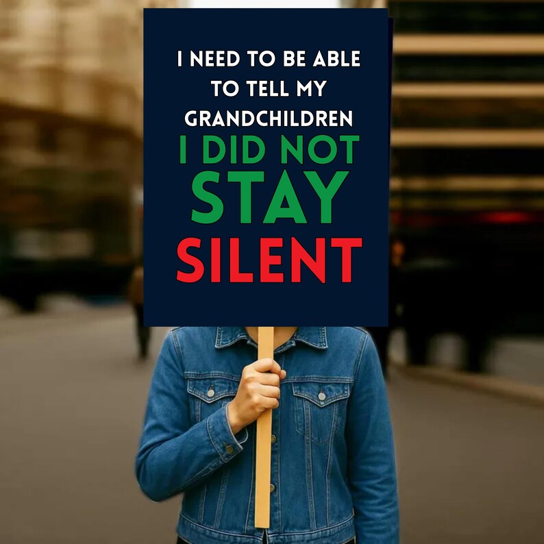 May include: A person holding a sign with the text "I NEED TO BE ABLE TO TELL MY GRANDCHILDREN I DID NOT STAY SILENT". The sign is dark blue with white, green, and red text. The person is wearing a denim jacket.