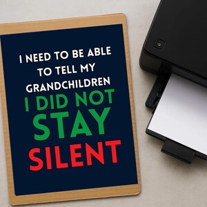 May include: A printed sign with the text "I NEED TO BE ABLE TO TELL MY GRANDCHILDREN I DID NOT STAY SILENT" in white, green, and red. A black printer is in the background, with a sheet of white paper being printed.
