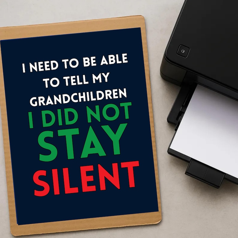 May include: A printed sign with the text "I NEED TO BE ABLE TO TELL MY GRANDCHILDREN I DID NOT STAY SILENT" in white, green, and red. A black printer is in the background, with a sheet of white paper being printed.