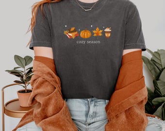 May include: Dark gray t-shirt with a fall-themed graphic. The design includes a pumpkin, maple leaf, and a cup of coffee. The words "cozy season" are printed below the illustrations. The person is wearing a rust-colored jacket and blue jeans.