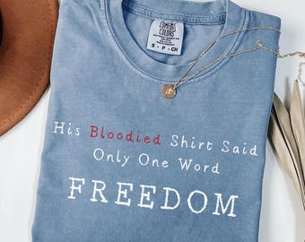 May include: A blue Comfort Colors t-shirt with the text "His Bloodied Shirt Said Only One Word FREEDOM" in white and red. A gold necklace with a pendant is draped across the shirt.