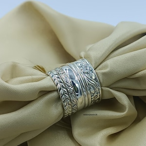 Peut inclure: Gros plan d'une bague en argent ornée de motifs floraux et de feuilles détaillés. La bague est composée de plusieurs anneaux, chacun avec un design unique. La bague est présentée sur un tissu beige, mettant en valeur ses détails.