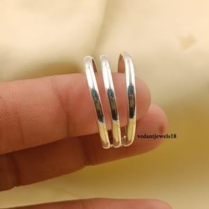 Puede incluir: Un anillo plateado con tres bandas. El anillo se muestra en un dedo sobre un fondo suave y neutro. El anillo tiene una superficie pulida y reflectante. El texto "vedantjewels18" es visible en la esquina inferior derecha.