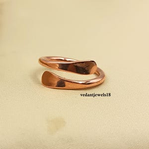 Puede incluir: Un anillo de color cobre con un diseño abierto. El anillo presenta dos extremos curvos de metal martillado que se superponen. El anillo se muestra sobre una superficie neutra y texturizada. El texto "vedantjewels18" es visible.