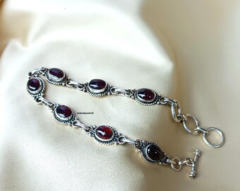 May include: A silver bracelet featuring oval-shaped, deep red gemstones. The gemstones are set in ornate, detailed silver settings. The bracelet has a toggle clasp and is displayed on a cream-colored surface.