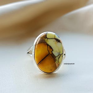 Peut inclure: Bague ovale en argent. La bague présente une pierre polie aux couleurs jaune, marron et crème, avec un motif craquelé unique. Le bijou est mis en valeur sur un fond neutre. Bijou de mode.