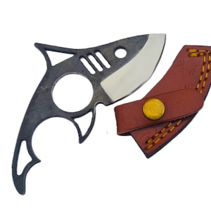 Può includere: Un coltello nero a forma di squalo con una lama curva e un fodero in pelle marrone. Il coltello ha un design unico con forme ritagliate e una lama argentata. Il fodero ha cuciture gialle e un bottone dorato.