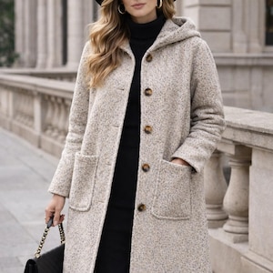 Può includere: Un cappotto lungo beige chiaro con cappuccio e ampie tasche. Il cappotto ha grandi bottoni marroni ed è indossato sopra un vestito nero. Un cappello e una borsa neri completano l'outfit. Il cappotto è un'elegante opzione di capospalla.