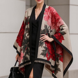 Op de afbeelding: Een beige sjaal met een groot bloemmotief in rood en roze, afgezet met zwart. De sjaal wordt over een zwarte jurk gedragen. Een zwarte handtas wordt in de linkerhand gehouden.