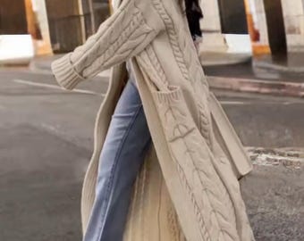 May include: A long, cream-colored cable-knit cardigan with a shawl collar and front pockets. The cardigan is knee-length and features a textured knit pattern. The person is wearing blue jeans and white shoes.