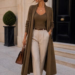 Pode incluir: Uma mulher vestindo um cardigan verde-oliva, top combinando e calças bege. Ela também usa óculos de sol, um colar de ouro e uma bolsa marrom. Um cinto e sapatos marrons completam o look. O fundo apresenta um edifício com uma porta preta.