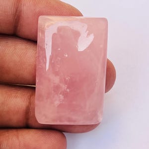 Puede incluir: Una piedra preciosa de cuarzo rosa rectangular con una superficie lisa y pulida de color rosa pálido translúcido. La piedra presenta inclusiones naturales y un acabado brillante que refleja la luz sobre su superficie plana y uniforme.