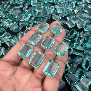 Puede incluir: Una colección de numerosas piedras preciosas sueltas, con corte esmeralda y un color azul verdoso translúcido. Las piedras presentan superficies facetadas que reflejan la luz, y varias de estas gemas rectangulares se muestran descansando sobre una palma abierta.