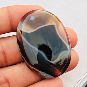 Può includere: Una pietra preziosa lucidata di forma ovale con una superficie liscia e brillante nei toni del marrone scuro e del nero. La pietra presenta distinti motivi a bande bianche e fluide che si curvano sul centro scuro, creando un aspetto stratificato.