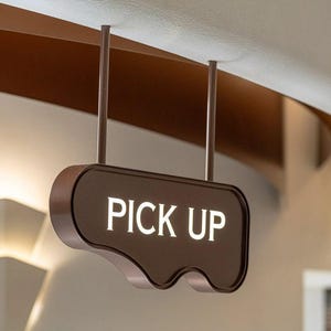 Op de afbeelding: Een bruin, verlicht "PICK UP" bord hangt aan het plafond. Het bord heeft een unieke, golvende onderrand en wordt opgehangen door twee dunne staven. Het bord bevindt zich in een moderne omgeving met muren in neutrale tinten en architecturale details.