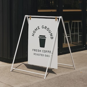 May include: A-frame sign with the words "HOME GROUND" and "FRESH COFFEE ROASTED DAILY" in black text. The sign features a dark green coffee cup graphic. The frame is white and stands on a concrete surface.