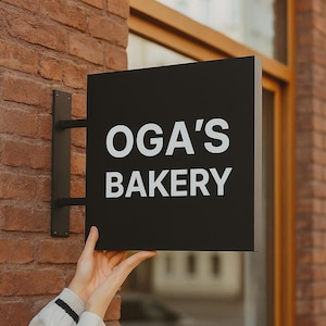 May include: A black square sign with the text "OGA'S BAKERY" in white lettering is mounted on a brick wall. The sign is held by a black metal bracket. The bakery sign is displayed outside a building.