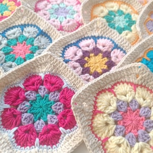 Könnte beinhalten: Nahaufnahme einer handgefertigten Häkeldecke mit mehreren bunten Blumenmustern. Jedes Quadrat hat einen cremefarbenen Rand und ein lebendiges, mehrfarbiges Blumenmuster in Rosa, Blau, Gelb und Lila. Die Decke ist aus weichem Garn.
