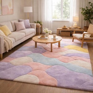 May include: A large, colorful area rug with a wavy, abstract design in pastel shades of pink, purple, yellow, and blue. The rug is placed in a living room setting with a sofa, coffee table, and natural light.