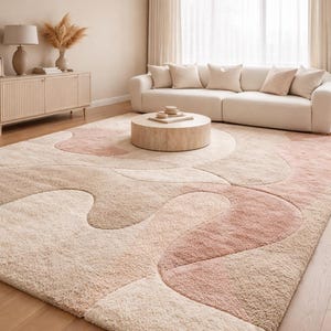 Op de afbeelding: Een groot, zacht vloerkleed met een abstract ontwerp in tinten beige, lichtbruin en roze. Het vloerkleed heeft een getextureerd oppervlak en een vloeiend, organisch patroon. Het staat in een woonkamer met een witte bank en een ronde salontafel.