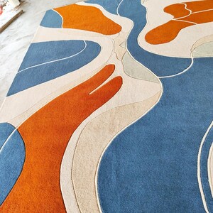 May include: A rectangular area rug featuring a tufted surface with an abstract pattern of flowing, organic shapes in shades of deep blue, bright orange, and cream. The design consists of curved lines and solid color blocks.