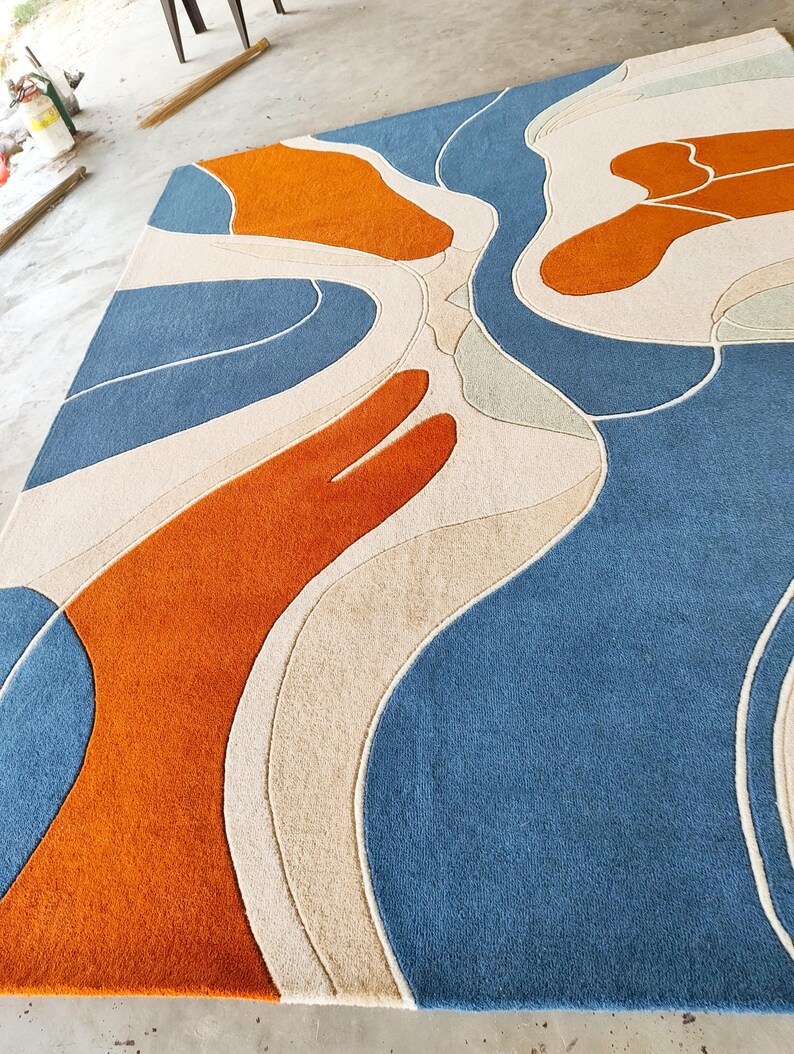 May include: A rectangular area rug featuring a tufted surface with an abstract pattern of flowing, organic shapes in shades of deep blue, bright orange, and cream. The design consists of curved lines and solid color blocks.