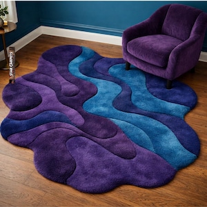 Op de afbeelding: Een zacht, abstract vloerkleed in tinten paars en blauw. Het vloerkleed heeft een golvende, organische vorm met kleurlagen, wat een modern en artistiek ontwerp creëert. Een paarse fauteuil staat in de buurt op een houten vloer.