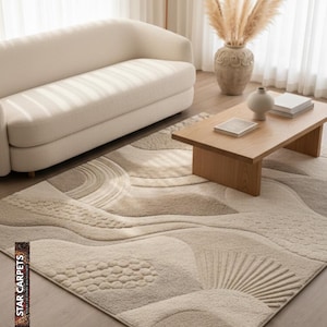 Op de afbeelding: Een beige vloerkleed met een textuur, abstract ontwerp met golfachtige patronen en geometrische vormen. Het kleed is geplaatst in een woonkamer met een crèmekleurige bank, houten salontafel en decoratieve vaas.