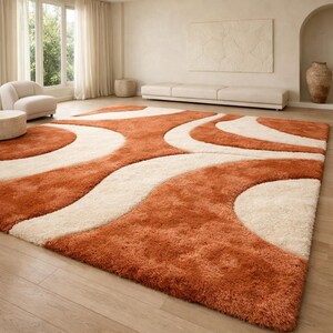 May include: A large, rectangular area rug with a plush, textured surface. The rug features a design of flowing, curved lines in shades of orange and cream. The rug is placed on a light-colored wooden floor in a well-lit room.