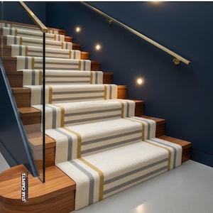 May include: A staircase with wooden steps and a cream-coloured carpet runner. The runner has grey and gold horizontal stripes. A glass panel and brass handrail are visible. The wall is painted a dark blue.