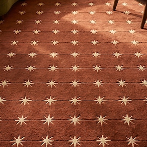 Op de afbeelding: Een rechthoekig vloerkleed in een diepe terracotta rode kleur met een herhalend rasterpatroon van crèmekleurige achtpuntige sterren. Het kleed heeft een getextureerd, getuft oppervlak met horizontale en verticale lijnen die vierkanten vormen.