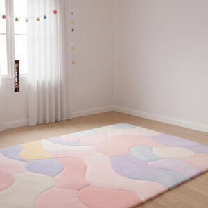 May include: A large, rectangular area rug with a pastel color palette of pink, purple, yellow, and blue. The rug features an abstract, flowing design, and is placed on a hardwood floor. The room has white walls and a window with sheer curtains.