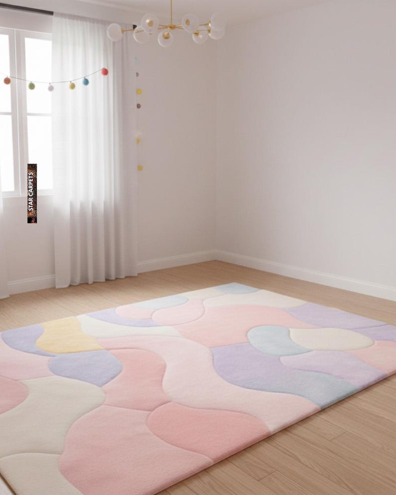May include: A large, rectangular area rug with a pastel color palette of pink, purple, yellow, and blue. The rug features an abstract, flowing design, and is placed on a hardwood floor. The room has white walls and a window with sheer curtains.
