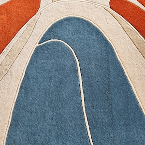 May include: A tufted area rug featuring a bold abstract pattern with large, curved shapes in deep blue, burnt orange, and cream. The design is defined by raised, carved lines that separate the distinct color blocks, creating a textured surface.