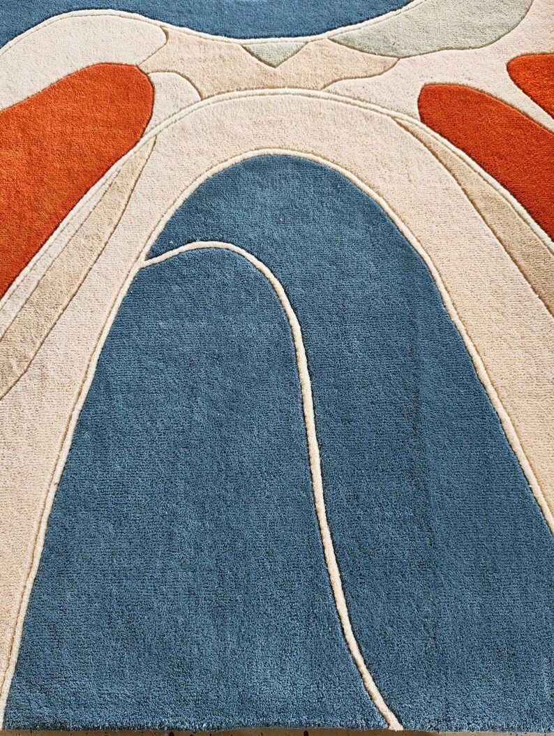 May include: A tufted area rug featuring a bold abstract pattern with large, curved shapes in deep blue, burnt orange, and cream. The design is defined by raised, carved lines that separate the distinct color blocks, creating a textured surface.
